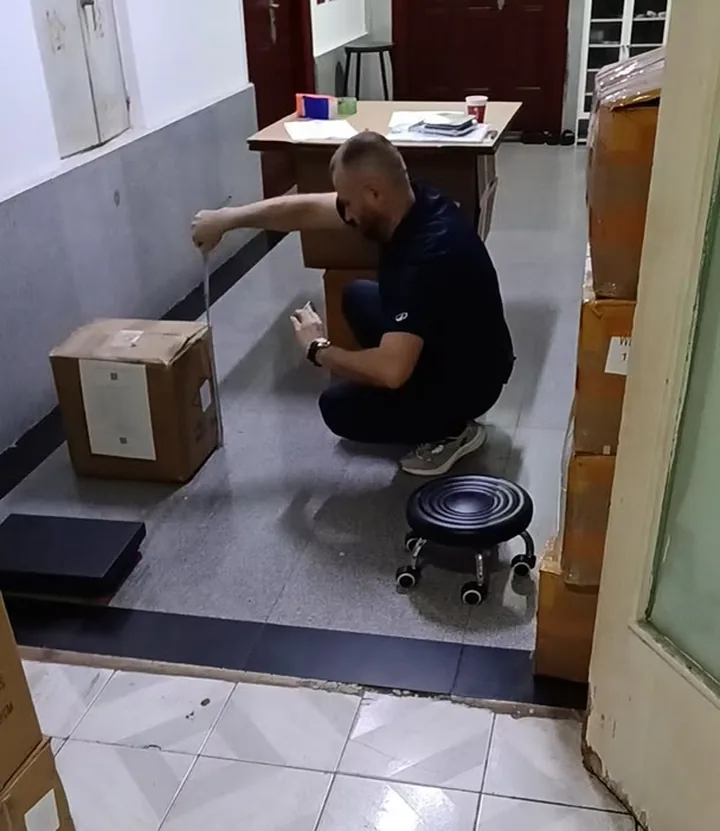 A real Qili Toys buyer visiting the Shenzhen warehouse in person, kneeling to seal his own brown export carton with packing tape, with additional cartons staged on either side and a paperwork desk behind him