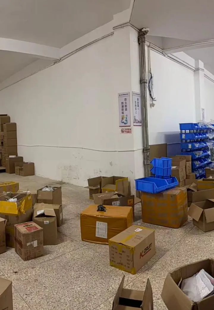 Qili Toys warehouse compliance corner with fire/damp/theft/rodent prevention and Safety First signage on the wall, organized blue parts-storage bins, and active mixed-SKU consolidation cartons in the foreground